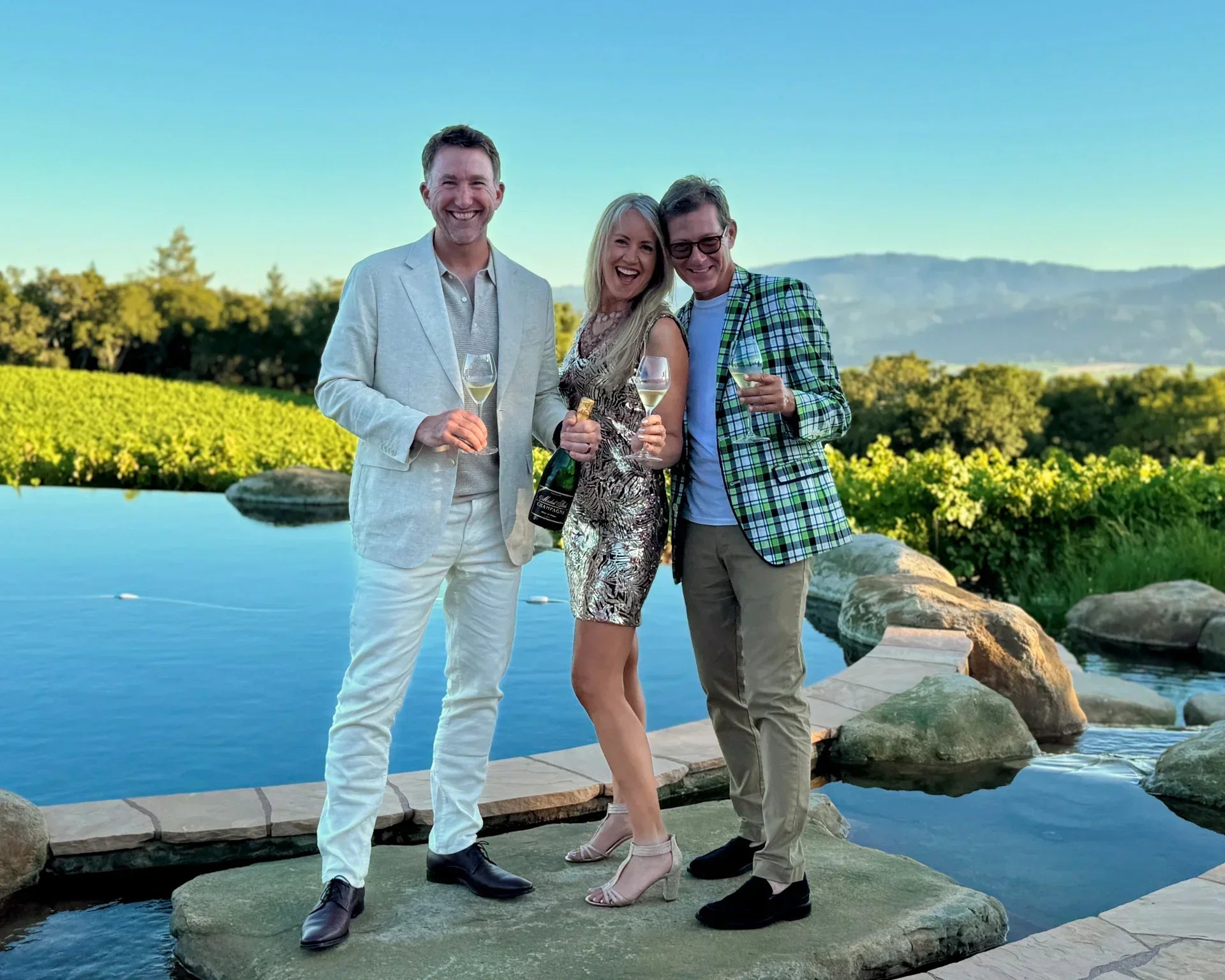 A group of friends posing in front of a pool and vineyards in the background holding a bottle of Michel Foch Champagne
