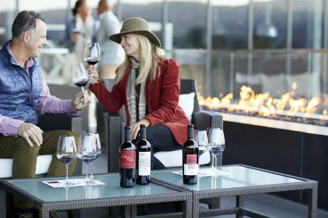A man and woman smiling at each other and drinking wine together outside on patio furniture by the fireplace