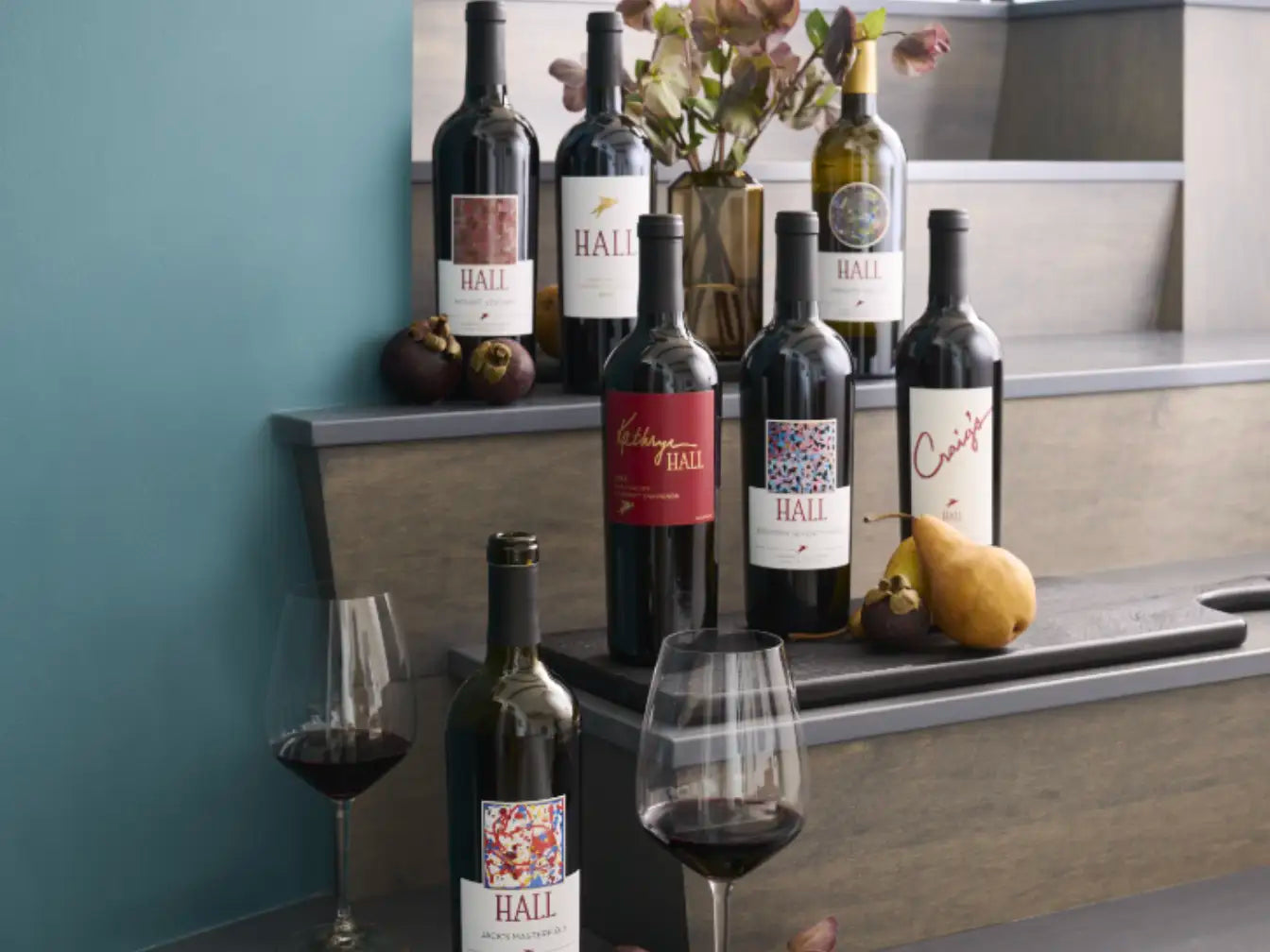 An assortment of HALL wines, including Kathryn Hall and Craig’s Cabernet Sauvignon, displayed on wooden tiers with fresh fruit and two glasses of red wine.