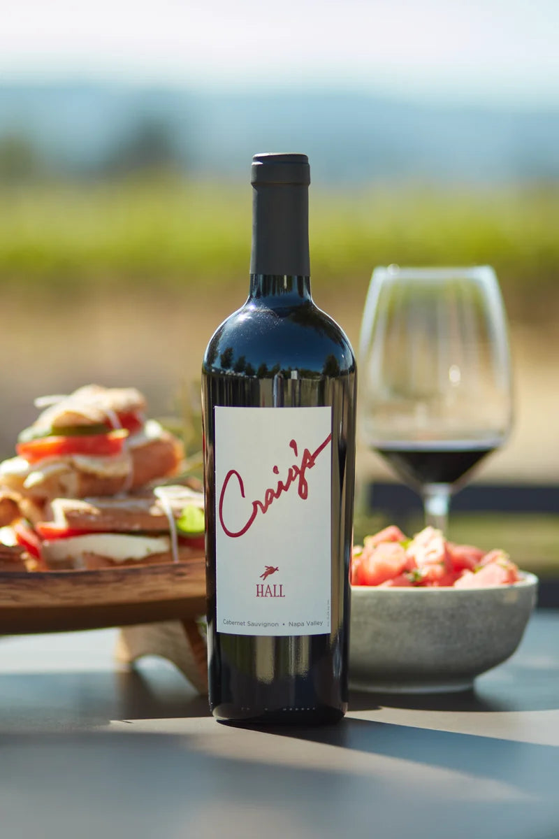 Photo of a bottle of 2019 HALL Craig's Cabernet Sauvignon on a picnic table with a platter of sandwiches, a summer salad, and glass of red wine in the background.