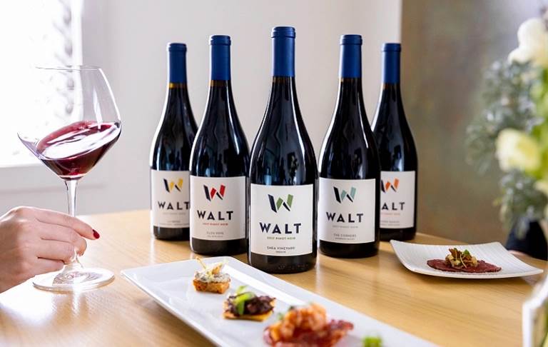 Wine bottles with 'WALT' label on a table with curated food pairings while a hand swirils a glass of Pinot Noir. 