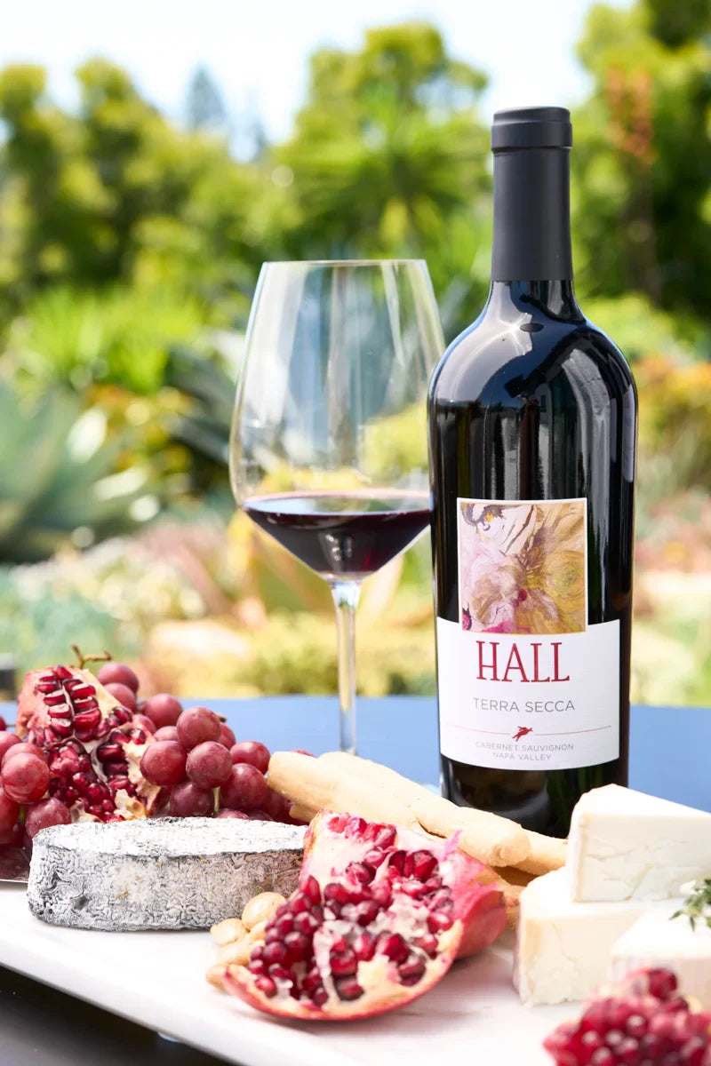 Bottle of 2022 HALL Terra Secca Cabernet Sauvignon on a table with a glass of red wine and a cheese platter in the foreground