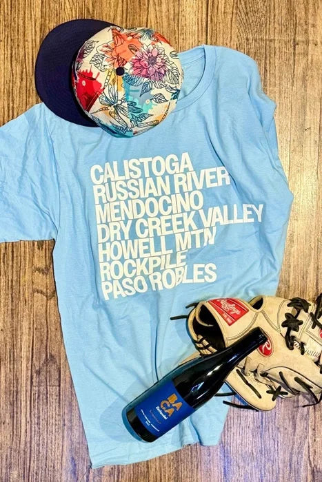 Flay lay image of a light blue BACA Wines t-shirt listing out all the names of the wine appellations used to produce their Zinfandel. The shirt is accompanied by a colorful baseball hat, bottle of BACA zinfandel and a baseball mitt.