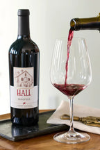 Bottle of HALL Bergfeld Cabernet Sauvignon on a wooden table beside red wine being poured into a glass, with a wine opener resting on a folded serviette.