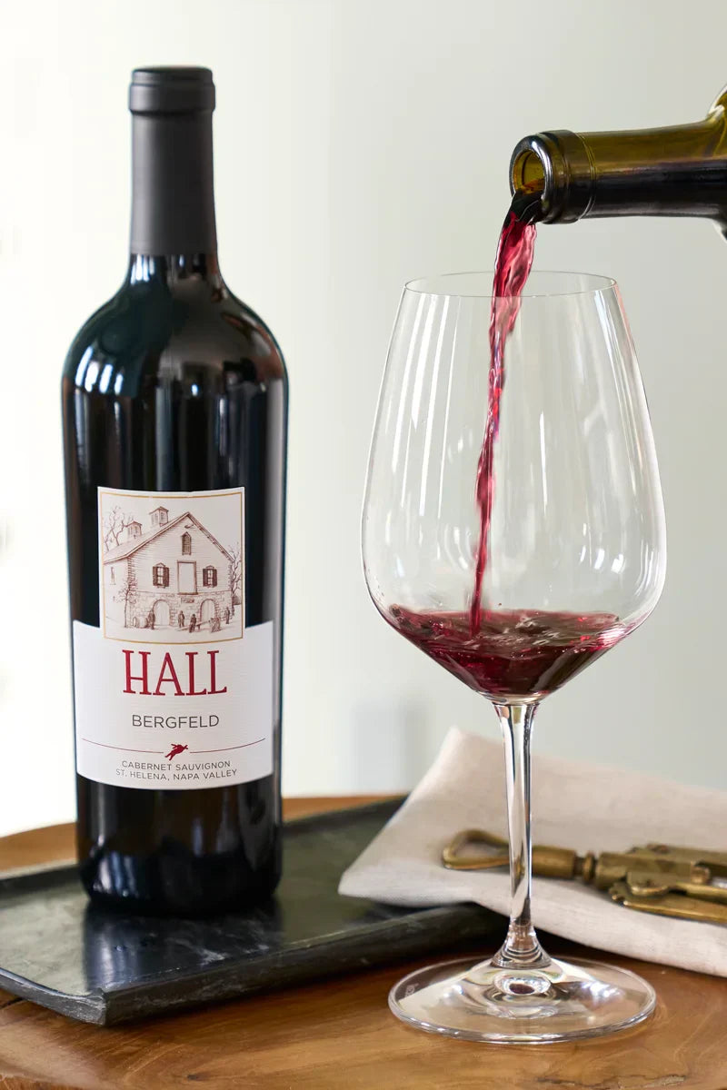 Bottle of HALL Bergfeld Cabernet Sauvignon on a wooden table beside red wine being poured into a glass, with a wine opener resting on a folded serviette.