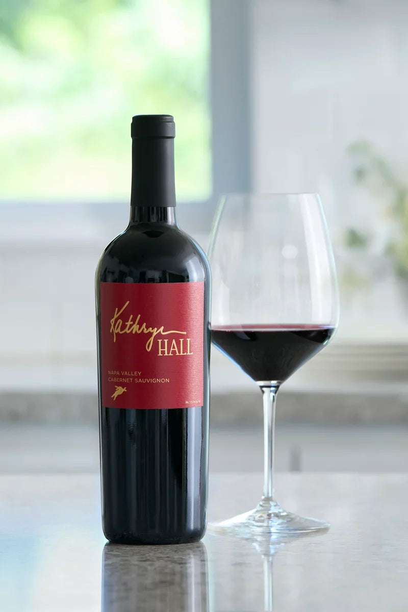 Image of a bottle of Kathryn Hall Napa Valley Cabernet Sauvignon nest to a glass of red wine displayed on a granite counter.
