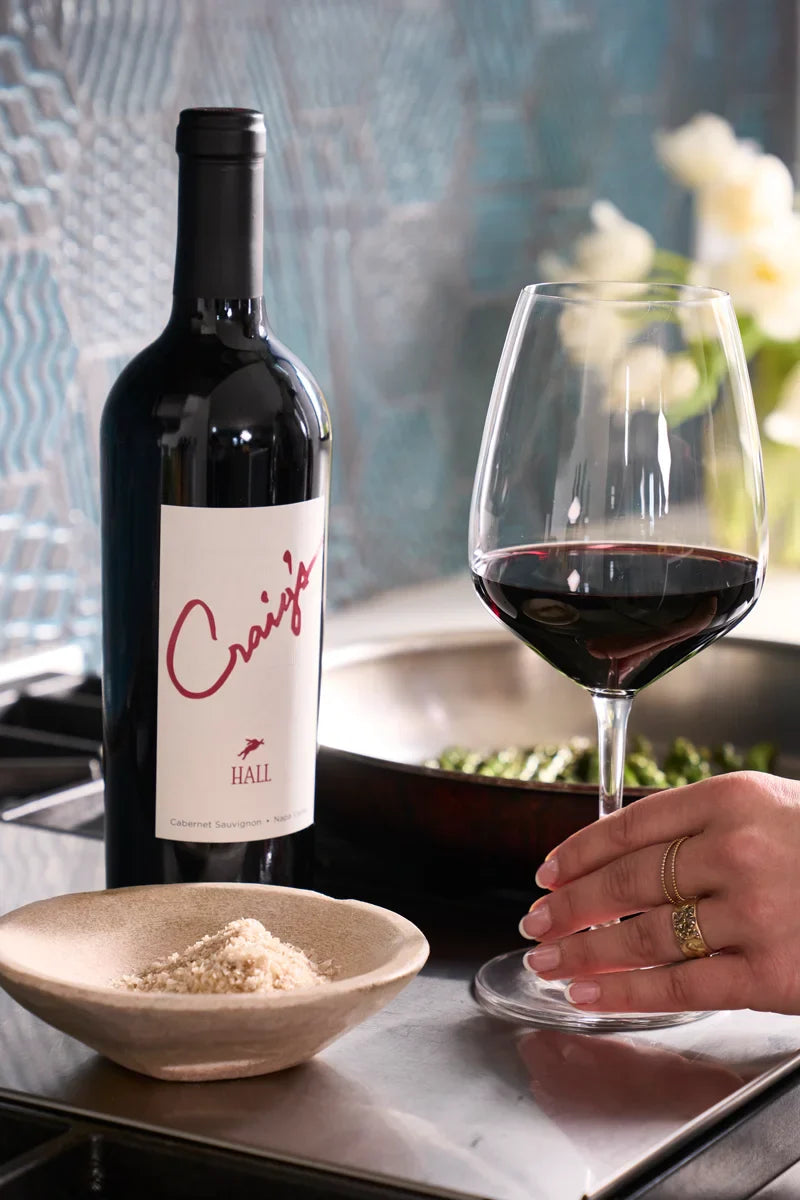 Woman reaching for a glass of red wine next to a bottle of 2019 HALL Craig's Cabernet Sauvignon, with a dish of shredded Parmesan and sautéing vegetables on the stove nearby