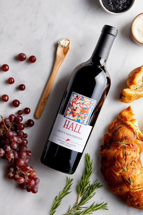 Wine bottle labeled 'Hall' with grapes, croissant, and herbs on a light surface