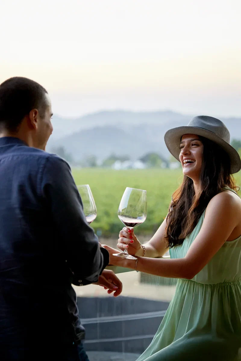 Couple smiling at each other while holding glasses of 2022 HALL The North End Cabernet Sauvignon at sunset, overlooking the HALL estate vineyard