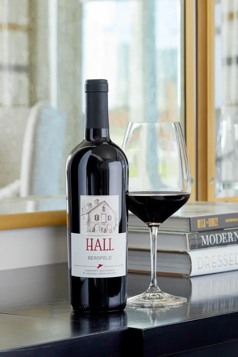 HALL Bergfeld Cabernet Sauvignon bottle on a console table beside a glass of red wine, with coffee table books and a window in the background