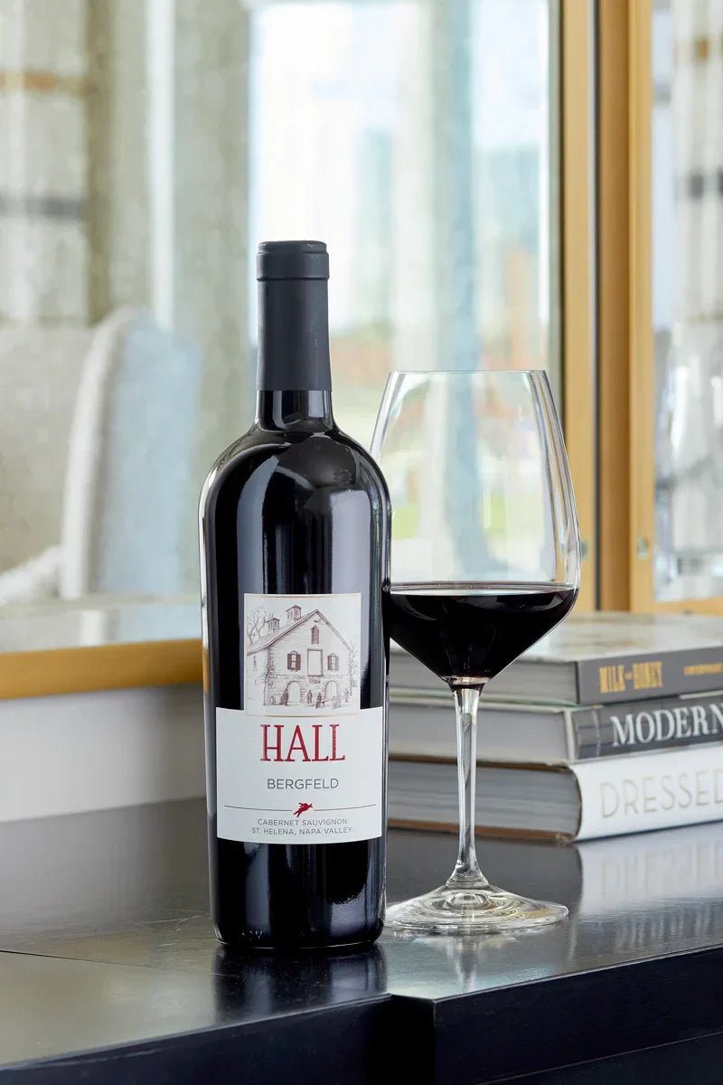 HALL Bergfeld Cabernet Sauvignon bottle on a console table beside a glass of red wine, with coffee table books and a window in the background