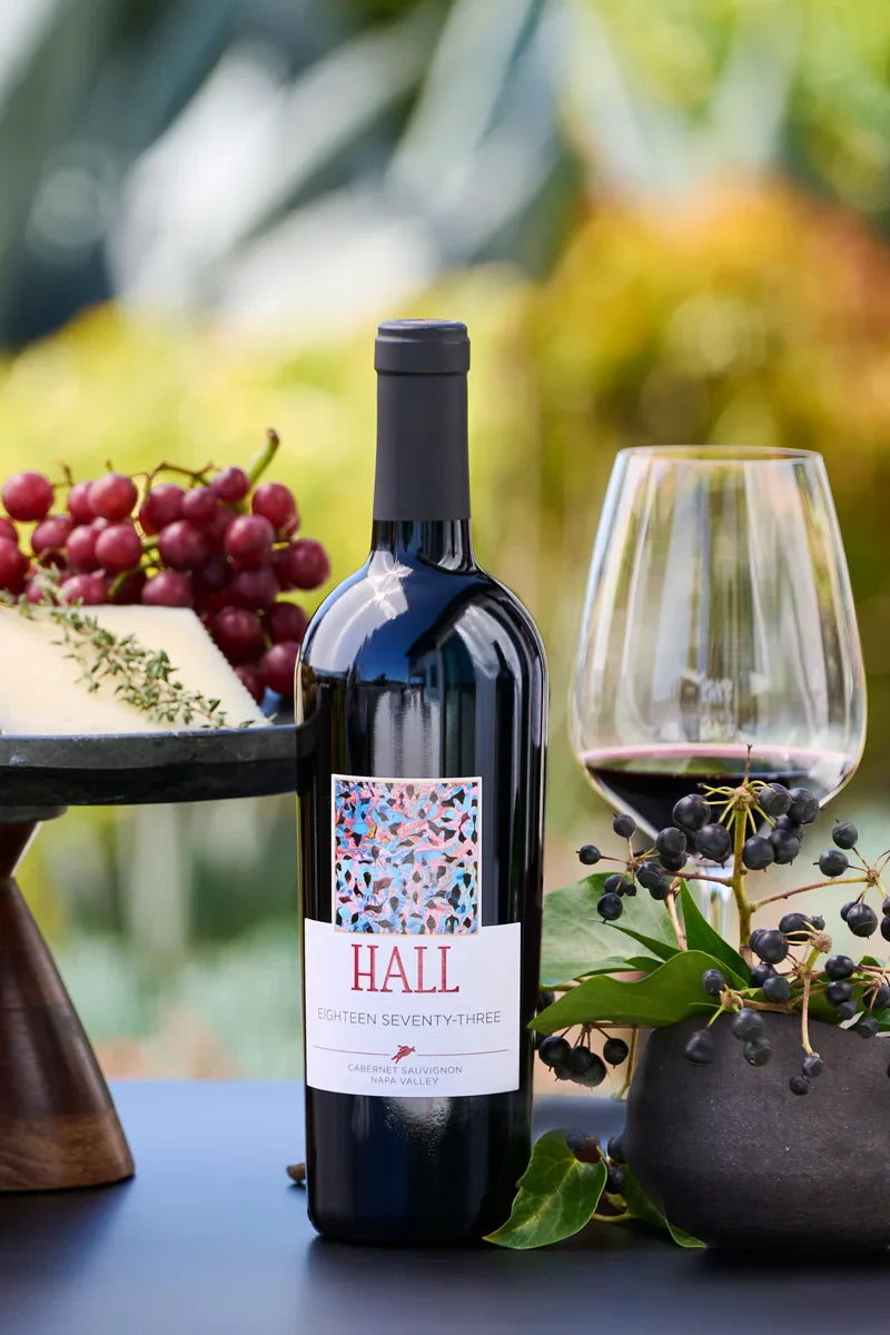 Bottle of 2021 HALL Eighteen Seventy-Three Cabernet Sauvignon beside a cheese and fruit platter and a glass of red wine
