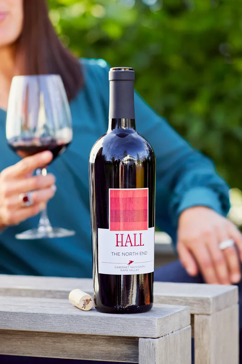 Woman seated in an outdoor wooden lounge chair holding a glass of red wine, with an opened bottle of 2022 HALL The North End Cabernet Sauvignon and its cork resting on the armrest