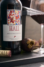Hall Jack's Masterpiece Cabernet Sauvignon wine bottle with a glass of wine and cork on a wooden surface.