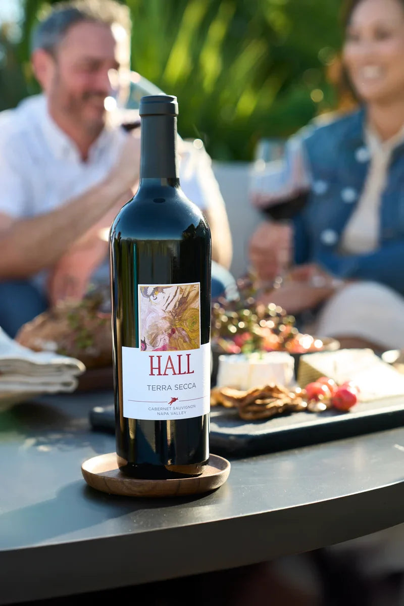 Bottle of 2022 HALL Terra Secca Cabernet Sauvignon on an outdoor lounge table, with a cheese platter and a couple enjoying red wine in the blurred background