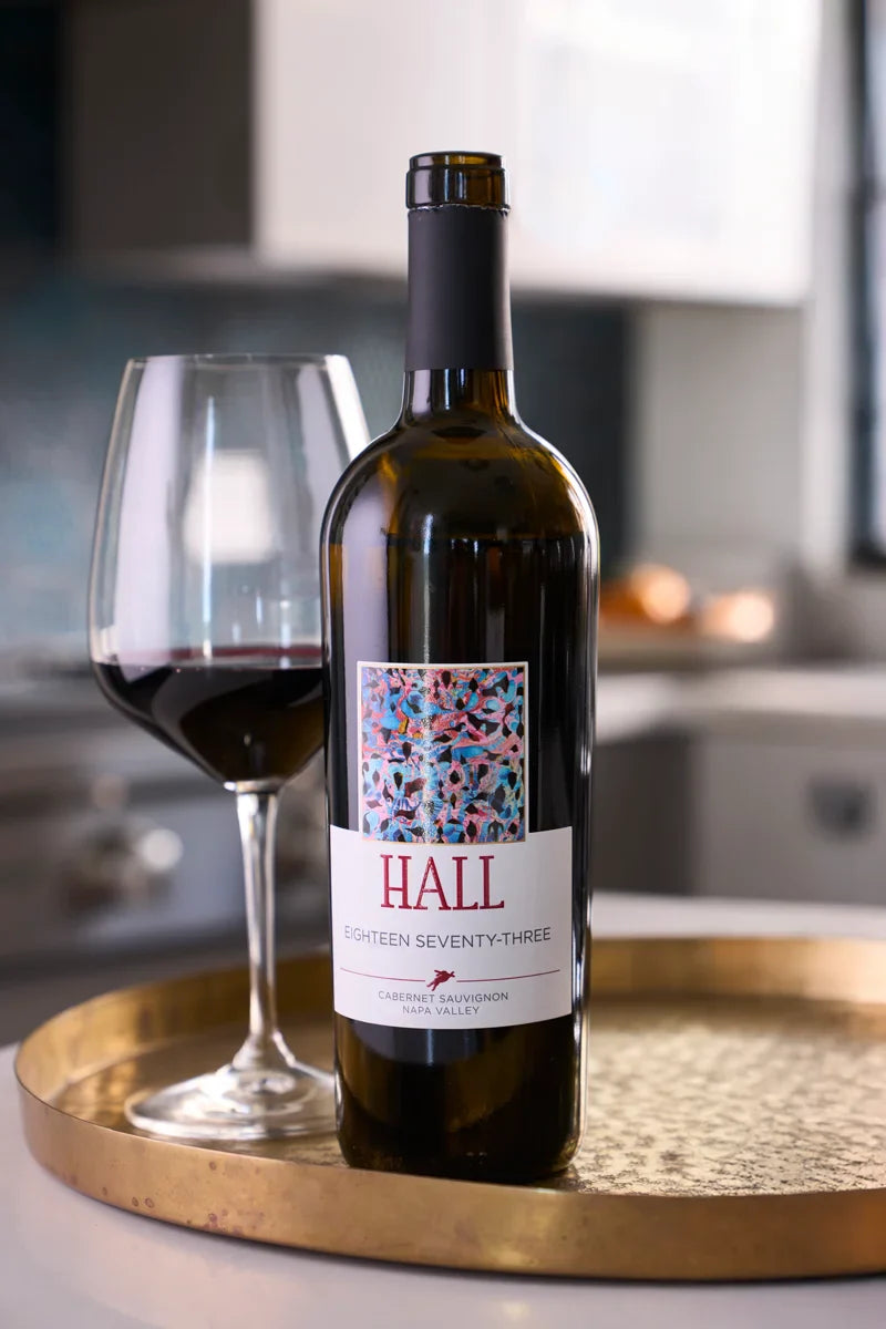 Bottle of 2021 HALL Eighteen Seventy-Three Cabernet Sauvignon on a tray next to a glass of red wine