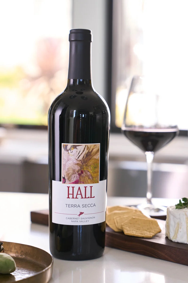 Bottle of 2022 HALL Terra Secca Cabernet Sauvignon on a kitchen counter, with cheese, crackers, and a glass of red wine on a wooden board in the background