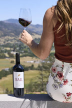 Woman seated on a ledge overlooking Napa Valley, holding a glass of red wine next to a bottle of HALL Napa Valley Cabernet Sauvignon.
