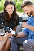 Couple enjoying outdoor wine tasting with 2021 HALL Ellie's Cabernet Sauvignon; man holding bottle and smelling glass of red wine