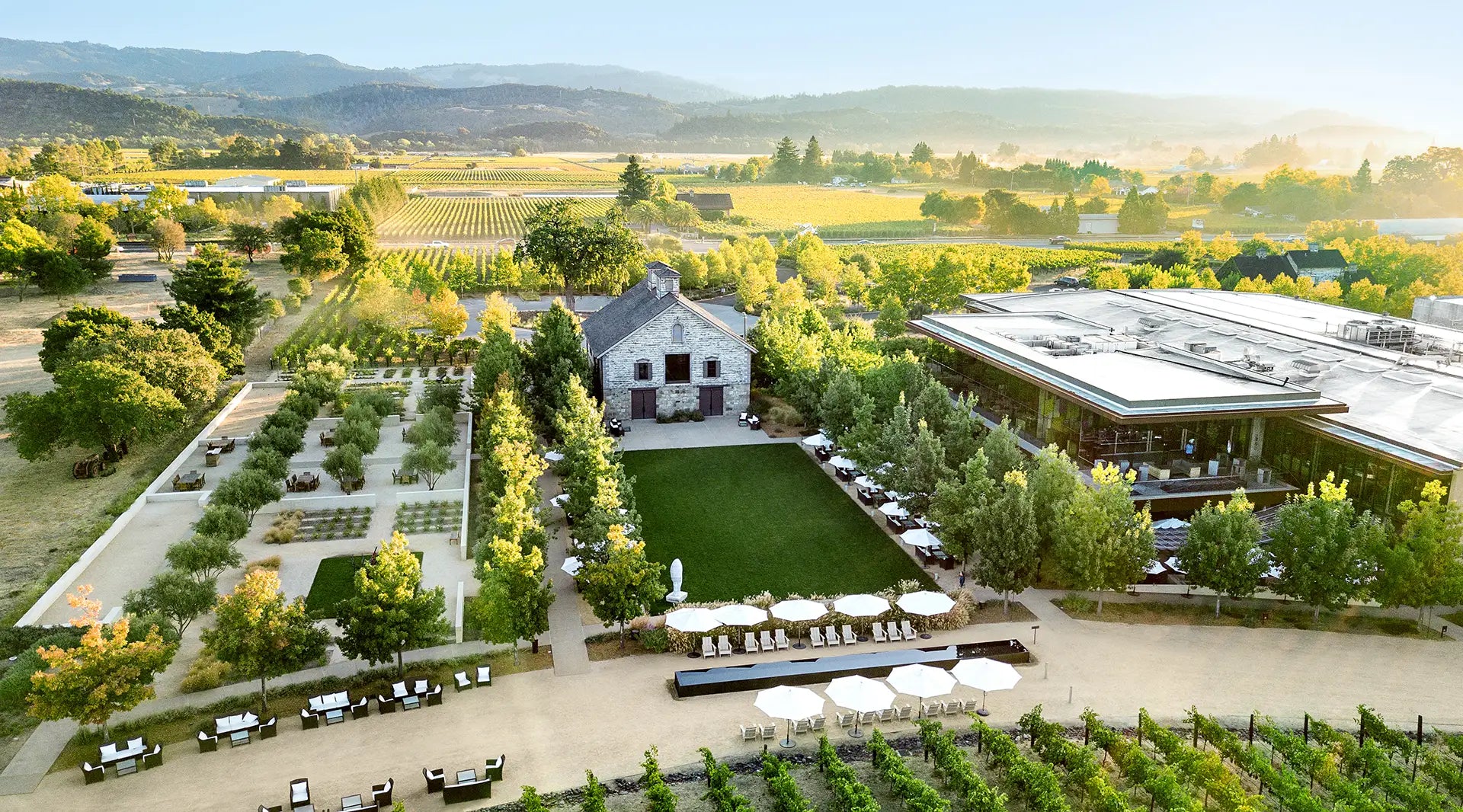 An aerial view of the HALL St. Helena estate at golden hour, featuring the historic stone Bergfeld Winery building, lush green lawns, and modern tasting facilities surrounded by rows of Napa Valley vineyards.