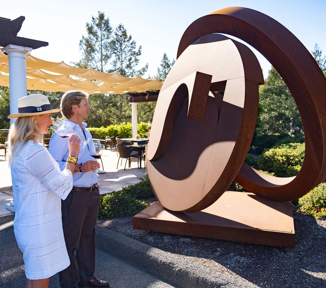 A couple enjoys glasses of red wine next to a large, modern rusted steel sculpture on the sun-drenched outdoor patio at HALL Rutherford in Napa Valley.