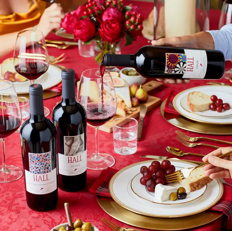 Red wine being poured from a HALL Coeur Cabernet Sauvignon bottle at a festive dinner table set with red roses, grapes, and cheese.