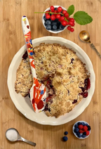 Image of a HALL Jack's Masterpiece rubber spatula resting in a berry crumble surrounded by fruit on a wood background.