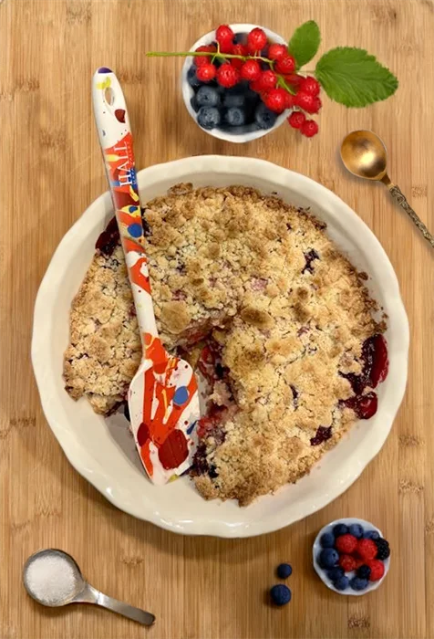 Image of a HALL Jack's Masterpiece rubber spatula resting in a berry crumble surrounded by fruit on a wood background.