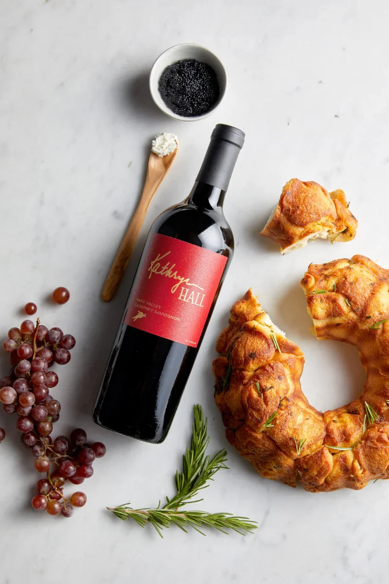 Image of a bottle of Kathryn Hall Napa Valley Cabernet Sauvignon laying flat on a marble countertop surrounded by herbed bread, caviar, red grapes and creme fraiche.