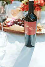 A bottle of Kathryn Hall Napa Valley Cabernet Sauvignon in a picnic setting with a cheese platter, flowers and a glass of red wine in the background.