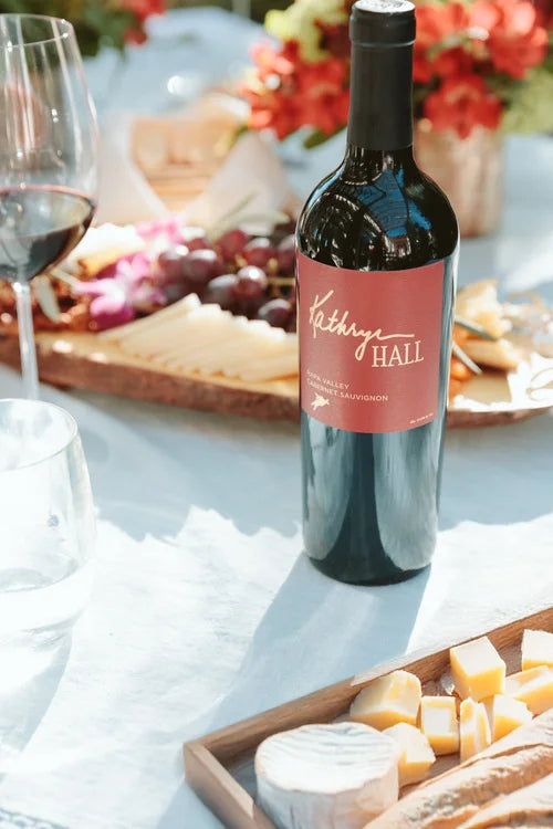 A bottle of Kathryn Hall Napa Valley Cabernet Sauvignon in a picnic setting with a cheese platter, flowers and a glass of red wine in the background.