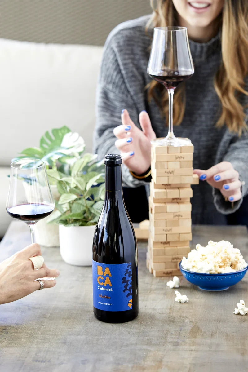 Two people enjoying wine and popcorn with a bottle of Baca wine on a table.