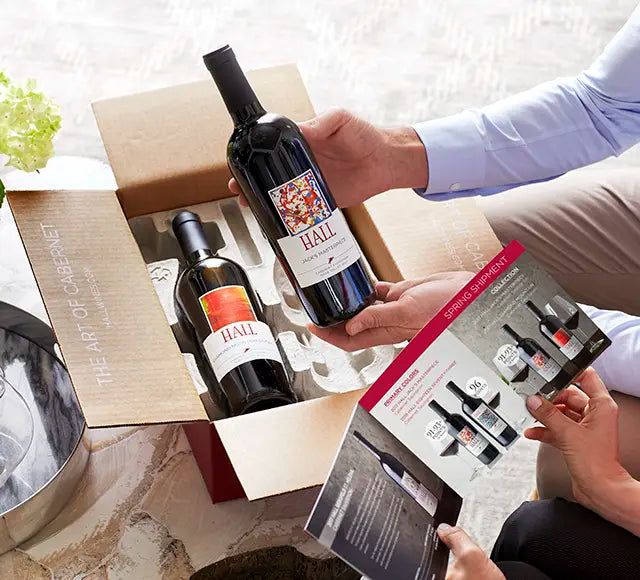 Two bottles of Hall Cabernet Sauvignon wine being held by a person with a box and leaflet in the background.