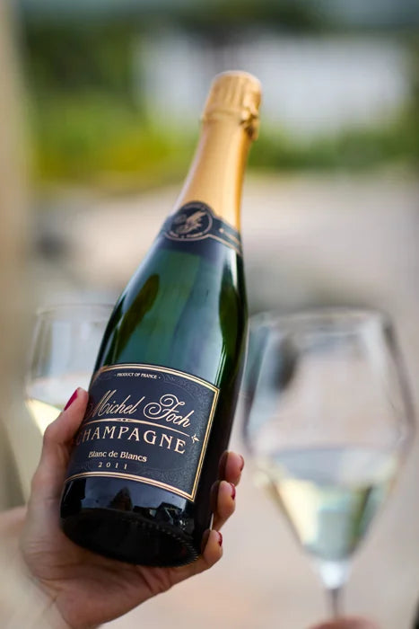 A hand holding a bottle of Michel Foch Blanc de Blancs Late Disgorged 2011 Vintage Champagne with champagne flutes in the background.