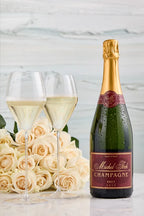 A bottle of Michel Foch Brut Champagne 2013 Vintage, two glasses of sparkling white wine, and a bouquet of white roses placed on a white marble table-top.
