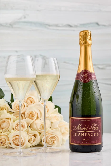 A bottle of Michel Foch Brut Champagne 2013 Vintage, two glasses of sparkling white wine, and a bouquet of white roses placed on a white marble table-top.