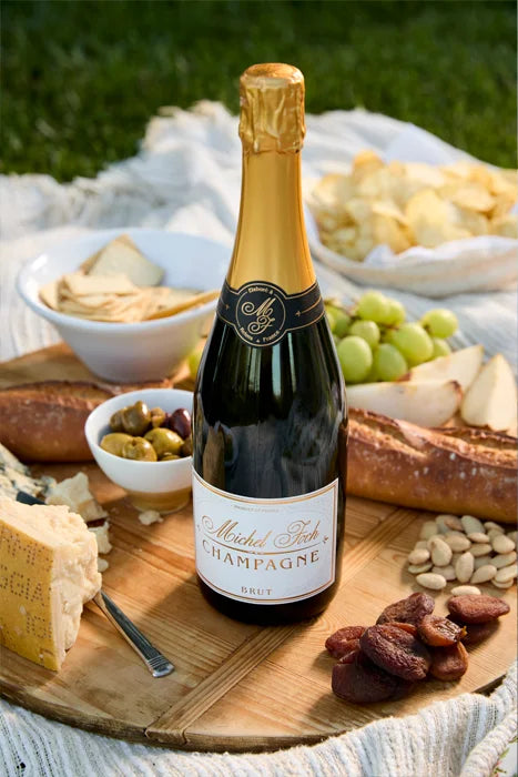 Bottle of Michel Foch Brut Champagne on a wood charcuterie board surrounded by cheese, olives, dried apricots, grapes, baguette, crackers and chips.