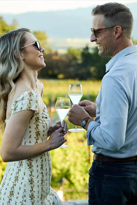 Couple enjoying Michel Foch Brut Champagne in a sunny vineyard, smiling at each other while holding glasses of sparkling wine.