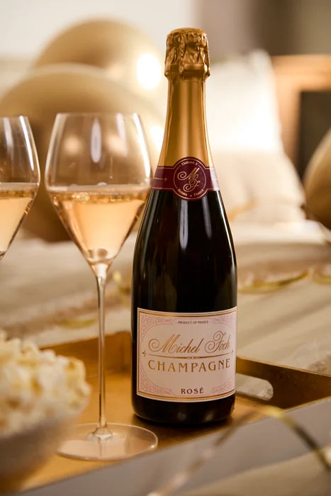 Festive scene with a bottle of Michel Foch Champagne Rosé and two glasses of sparkling rosé wine on a gold tray, gold balloons in the background, and a bowl of popcorn partially visible on the left.