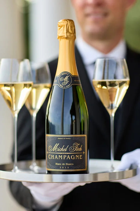 A man holding a tray with a bottle of Michel Foch Blanc de Blancs 2015 Vintage Champagne and 3 filled champagne flutes.