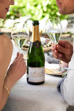 Man and woman smiling at each other while holding glasses of sparkling white wine, with a bottle of Michel Foch Brut Champagne on the table in front of them.