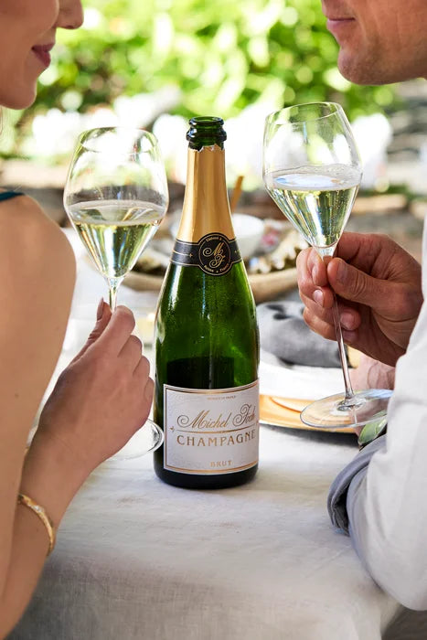 Man and woman smiling at each other while holding glasses of sparkling white wine, with a bottle of Michel Foch Brut Champagne on the table in front of them.