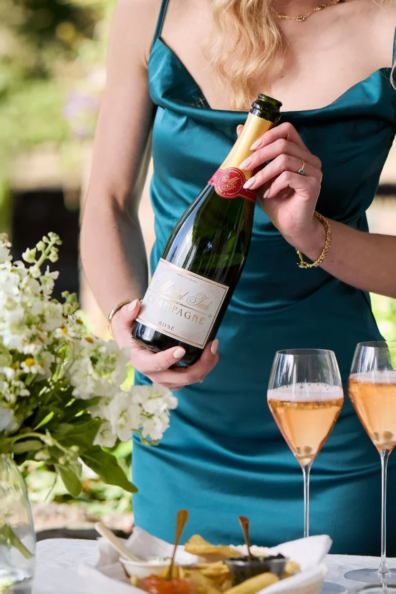A woman in an elegant green dress holding a bottle of Michel Foch Rose Champagne standing behind a table with two fluted full of Champagne and a tray of caviar.