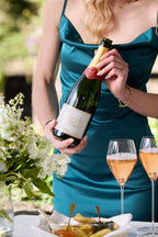 A woman in an elegant green dress holding a bottle of Michel Foch Rose Champagne standing behind a table with two fluted full of Champagne and a tray of caviar.