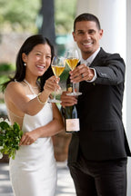 A bride and groom clinking their glasses of Michel Foch Brut Champagne. The groom holds a bottle of Michel Foch Brut Champagne in his free hand while the bride holds a bouquet of flowers.