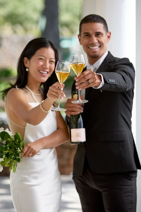 A bride and groom clinking their glasses of Michel Foch Brut Champagne. The groom holds a bottle of Michel Foch Brut Champagne in his free hand while the bride holds a bouquet of flowers.
