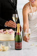 Bride and groom reaching for full champagne flutes on a table with a wedding cake and a bottle of Michel Foch Brut Champagne 2013 Vintage.