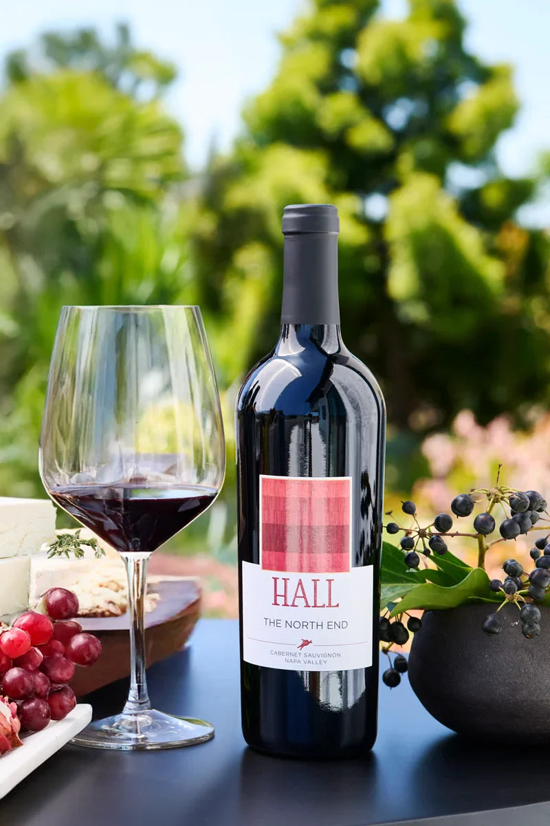 Bottle of 2022 HALL The North End Cabernet Sauvignon on an outdoor table beside a glass of red wine and a cheese and fruit plate