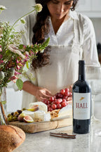 Woman in a kitchen creating a cheese board with figs, nuts, and red grapes. A bottle of HALL Napa Valley Cabernet is in the foreground next to a wine glass.