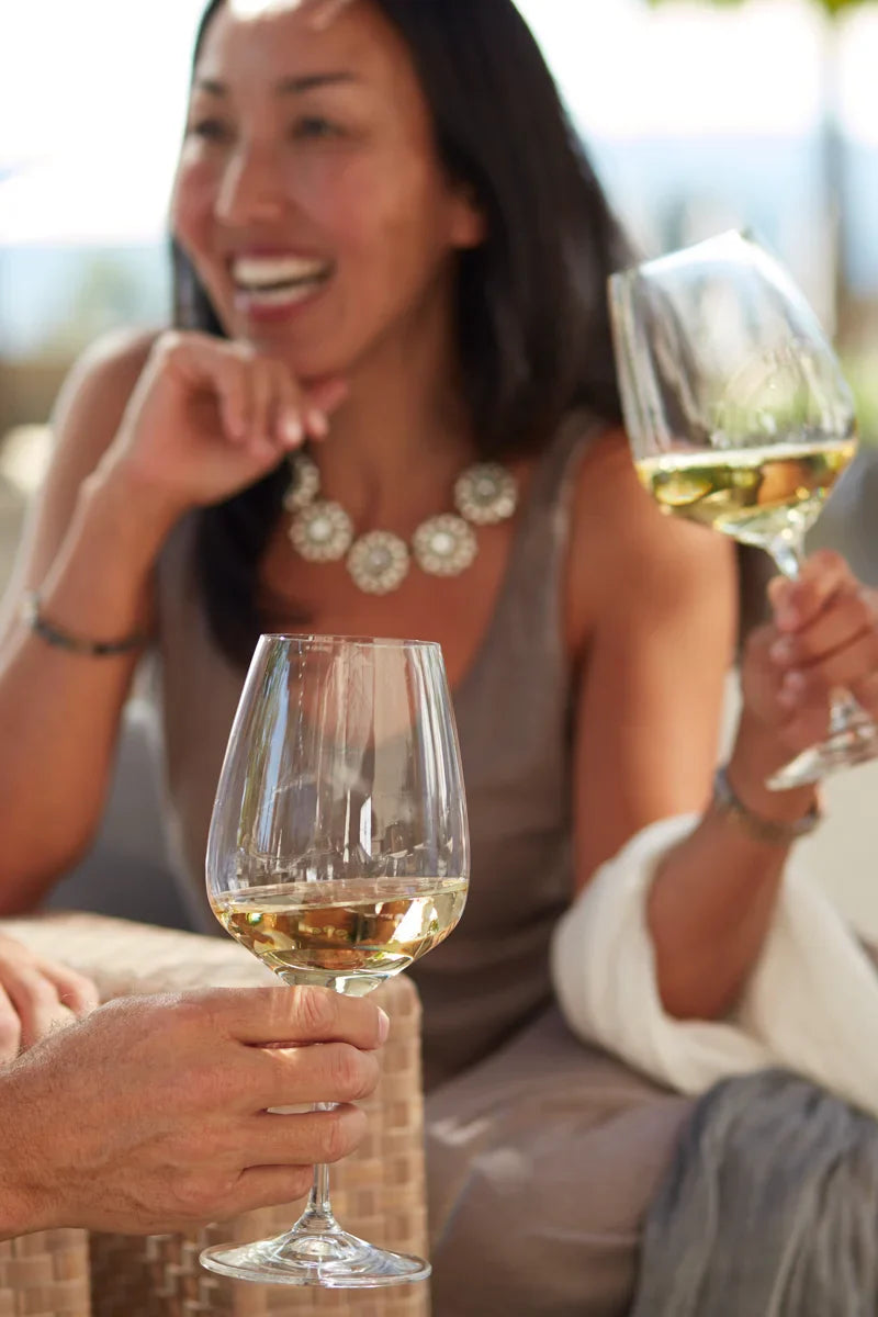 Image of a person holding a glass of 2024 HALL Napa Valley Sauvignon Blanc. In the blurred background, a woman is smiling and holding a glass of white wine.